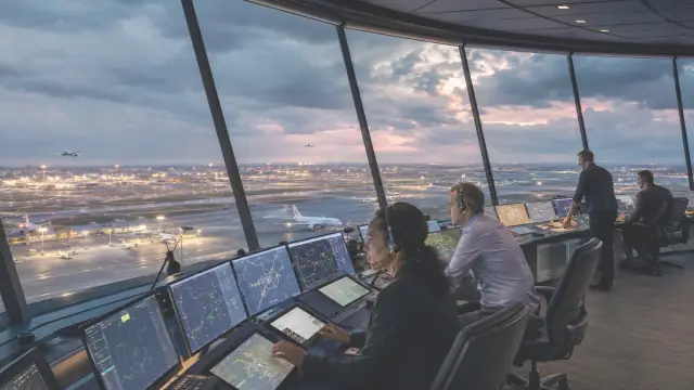 Reconstruction of a control tower cab at dusk, controllers at their displays with the airfield's taxiways and approach lights visible through the glass.