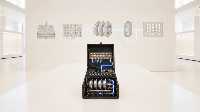 Studio reconstruction of an Enigma machine open on a white ground, rotors and plugboard cabling visible, with separate rotors displayed behind it.