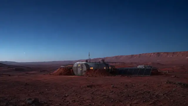 Speculative rendering of a small, partially buried crewed habitat with one lit window and a solar array on a rocky Martian plain beneath a darkening twilight sky.