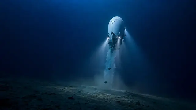 Reconstruction of a crewed deep-sea submersible suspended vertically in dark blue water above the abyssal seafloor, floodlights lit and a plume of released ballast falling beneath its hull.