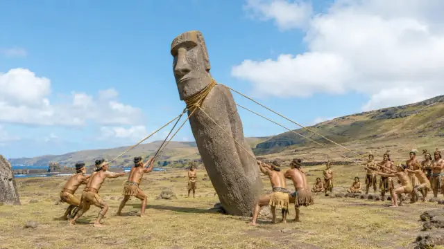 Reconstruction of an upright moai being walked with ropes by a team of people across open Rapa Nui grassland.