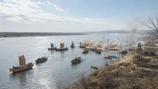 Reconstruction of the Red Cliffs engagement: linked wooden warships burning on the Yangtze at dusk, smoke rolling downriver past the cliffs.